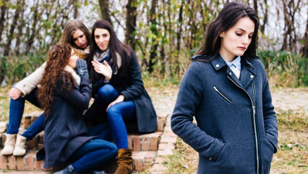 Sad-girl-with-friends-gossiping-in-background-behind-her-back-shutterstock