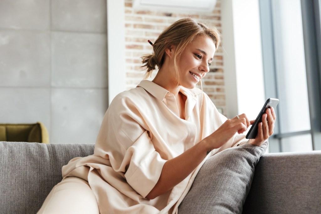 młoda kobieta siedzi uśmiechnięta w mieszkaniu na kanapie i korzysta ze smartfona