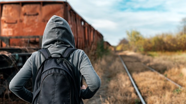 Młoda osoba w kapturze i plecaku idzie po opuszczonych torach kolejowych