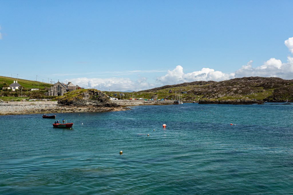 Boats-and-buoys-in-the-bay-the-rocky-shoreline-with-its-small-harbor-its-church-on-Inishbofin-Island-sunny-spring-day-with-blue-sky-in-County-Galway-Ireland-.jpeg