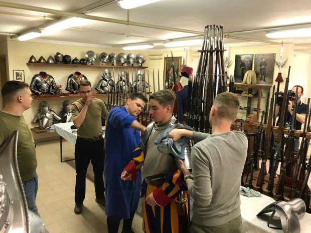 Young guards help their comrade to don his cuirass.