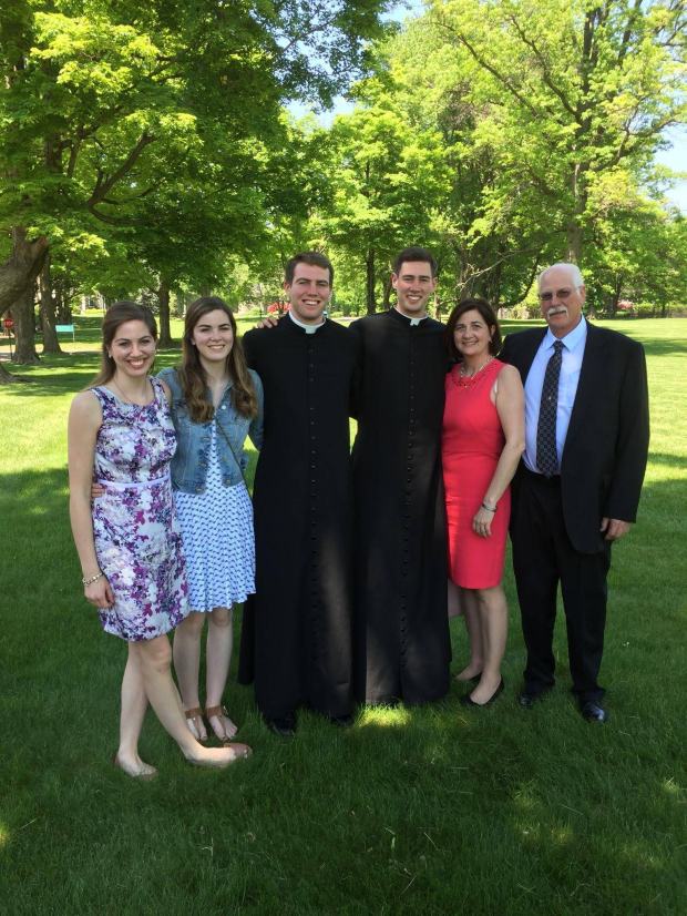 Family posing outside. Two sons in the middle in cassocks.
