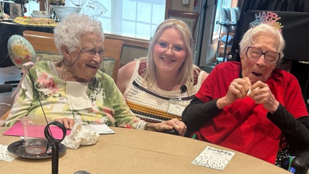 two elderly women with a blonde younger woman in the middle. one has a happy birthday hat