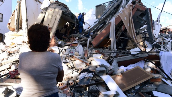 A woman looks at rescuers searching for survivors amid the rubble of a collapsed building after an earthquake in Pereira, Colombia on August 10, 2026. A magnitude 7.4 earthquake struck Colombia on August 10, 2026, and killed at least 111 people, damaged more than 1,500 buildings, and left an unknown number of people buried under rubble. The government declared a state of emergency to attend to the quake that shook several cities across the coffee-producing region, the Pacific coast, and neighboring Latin American countries, sending terrified residents racing into the street.